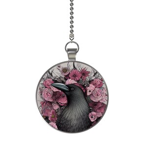 May include: A silver chain with a round pendant featuring a black crow surrounded by pink flowers. The crow has a black beak and black eyes. The flowers are drawn in a detailed style.