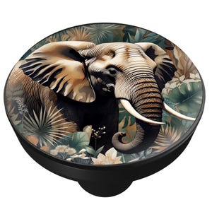May include: A black round phone grip featuring an illustration of an elephant with large tusks and detailed skin. The elephant is surrounded by lush green and brown foliage. The design is set against a black background.