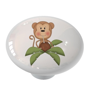 May include: A ceramic knob with a cartoon monkey drinking from a coconut. The monkey is brown with a light brown face and is surrounded by green palm leaves.