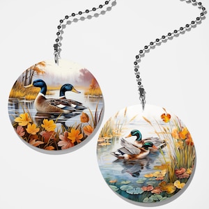 May include: Two round fan pull ornaments with images of ducks swimming in a pond with fall foliage. The ducks are brown, green, and blue with orange beaks. The pond is blue with green reeds and lily pads. The foliage is orange, yellow, and brown.