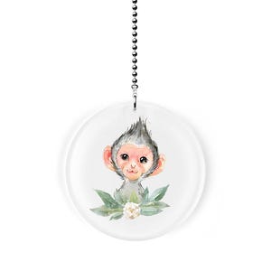 May include: A round, clear glass ceiling fan pull featuring a watercolor illustration of a monkey with gray fur and pink ears. The monkey is surrounded by green leaves and a white flower. A silver chain hangs from the top.