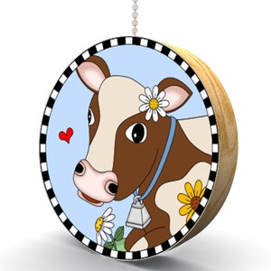 May include: A round wooden pull chain with a blue background and a cartoon cow with a white and brown spotted pattern. The cow has a blue collar with a bell and a daisy in its hair. There are other daisies around the cow.