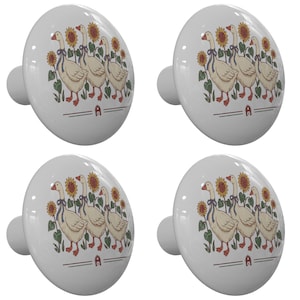 May include: Four white ceramic cabinet knobs. Each knob features a design of geese with sunflowers and the letter "A". The geese are cream-colored with blue bows. The sunflowers are yellow and brown.