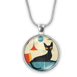 May include: Silver-tone pendant necklace featuring a round charm with a retro illustration of a black cat lounging on a blue surface. The background includes colorful hanging lamps and a cream-colored wall.