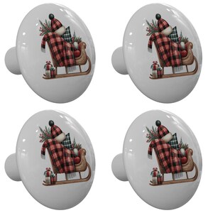 May include: Four white ceramic cabinet knobs with a Christmas theme. Each knob features a sleigh with a red and black plaid blanket, a hat, and gifts. The knobs have a silver-colored stem.