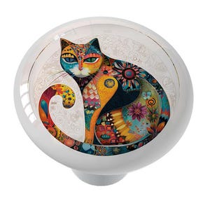 May include: A white ceramic cabinet knob featuring a colorful, stylized cat design. The cat is depicted with intricate patterns in shades of blue, orange, yellow, and purple. The knob has a glossy finish and a decorative, floral-patterned background.