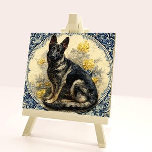 May include: A decorative tile featuring a German Shepherd dog illustration. The dog is depicted in black and gray, set against a backdrop of yellow flowers and a cream-colored background. The tile is framed with a blue and white patterned border, displayed on a small easel.