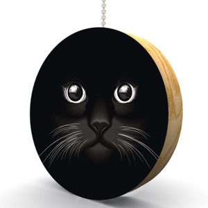 May include: A black cat's face with white whiskers peeks out from a dark background. The cat's eyes are large and round, and its nose is small and pink. The image is printed on a round wooden pull chain.