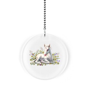 May include: A white unicorn foal lying in a field of white and purple wildflowers. The foal has a pink nose and a pink horn. The image is on a round, clear acrylic fan pull.
