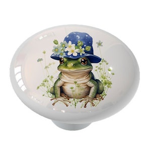 May include: A green frog wearing a blue and white polka dot hat with white flowers. The frog is sitting on a bed of green leaves.