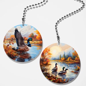May include: Two round fan pull ornaments with a fall theme. One features a single duck swimming in a lake with fall foliage in the background. The other features two ducks swimming in a lake with a sunset and fall foliage in the background.