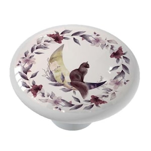 May include: A ceramic knob with a black cat sitting on a crescent moon surrounded by a wreath of purple and pink flowers.