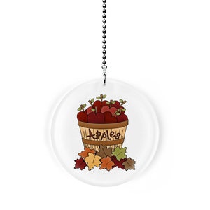May include: A round, white, decorative pull chain with a brown basket filled with red apples and the word "Apples" written on the side. The basket is surrounded by brown, orange, and green leaves.