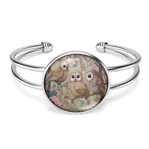 May include: Silver bangle bracelet with a round glass cabochon featuring a whimsical illustration of three owls perched on branches with pink flowers. The owls are brown and beige with detailed feathers and large eyes.