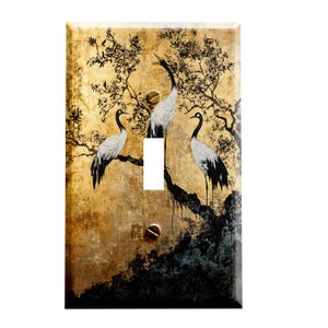 May include: A light switch cover with a gold background and a black and white illustration of three cranes perched on a tree branch. The background has a distressed, aged look.