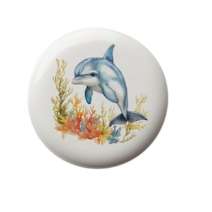May include: A white, round ceramic coaster featuring a watercolor illustration of a blue and white dolphin. The dolphin is surrounded by colorful coral and seaweed. The coaster has a glossy finish.