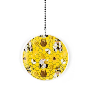 May include: A round, white ceiling fan pull with a yellow and black honeycomb pattern. The pattern features gnomes, honey pots, and sunflowers.
