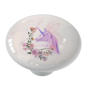 May include: A white ceramic knob with a purple unicorn with pink wings and flowers. The unicorn is surrounded by pink and purple flowers.