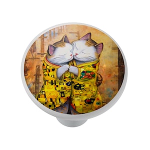 May include: A white cabinet knob featuring an illustration of two cats embracing. The cats are wearing yellow robes with geometric patterns. The background is a warm, textured design.