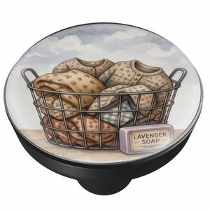 May include: A round decorative knob featuring a circular illustration of a wire laundry basket filled with folded tan patterned clothing. A rectangular bar of soap labeled LAVENDER SOAP sits in front of the basket against a cloudy sky background.