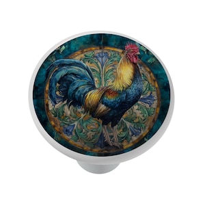 May include: A white cabinet knob featuring a colorful rooster illustration. The rooster has blue, yellow, and red plumage, set against a teal and gold background with floral accents. The knob is a decorative hardware element.