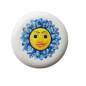 May include: A white ceramic button with a blue and yellow sun face design. The sun has a smiling face with blue eyes and a yellow background. The sun is surrounded by blue and white floral designs.
