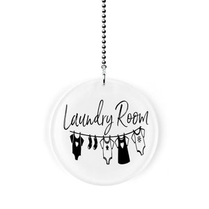 May include: A white round acrylic sign with a black chain hanging from the top. The sign says "Laundry Room" in black cursive writing with a line of black clothes hanging below the text.