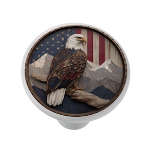 May include: A white cabinet knob with a detailed wooden carving of a bald eagle perched on a branch. The background features the American flag and mountains. The eagle has brown, white, and red feathers.