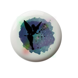 May include: A white ceramic button with a silhouette of a fairy with wings and a wand, surrounded by a watercolor design of blue, green, and purple with white stars.