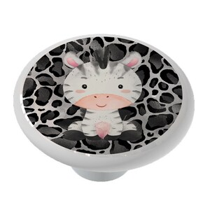 May include: A white ceramic knob with a black and white zebra print background. A cartoon zebra with black stripes and pink cheeks is in the center of the knob.