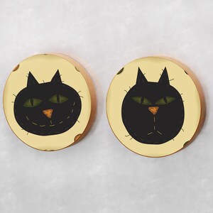 May include: Two round wooden wall decor pieces with a black cat face design on a cream background. The cat has green eyes and a stitched-on orange nose.