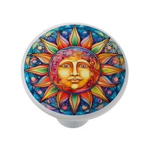 May include: A white cabinet knob with a colorful sun design. The sun features a smiling face with orange, yellow, and pink hues, surrounded by a stained-glass style pattern in blue, green, and orange. The knob is round.