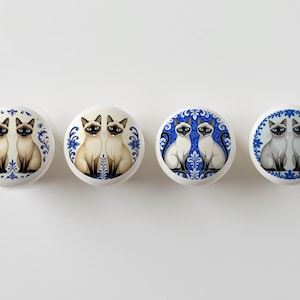 May include: Four white ceramic cabinet knobs with Siamese cat illustrations. Each knob features a pair of cats with blue eyes, set against a blue and white decorative border. The knobs are round and have a smooth finish.