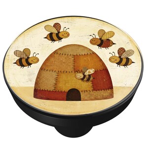 May include: A round, black PopSocket with a whimsical design featuring a beehive and several cartoon bees. The beehive is made of patchwork fabric in shades of brown and tan. The bees have brown and yellow striped bodies and patterned wings.