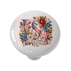 May include: A white ceramic cabinet knob with a patriotic design. The word "USA" is stylized with the American flag, surrounded by red, white, and blue flowers. A banner reads "250 YEARS OF FREEDOM".