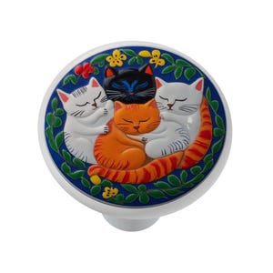 May include: A round ceramic cabinet knob featuring a raised, colorful illustration of four sleeping cats in black, orange, and white. The cats are nestled together against a deep blue background, surrounded by a border of green leaves and yellow flowers.