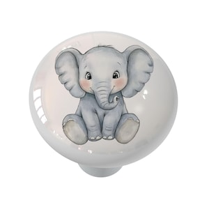May include: A white ceramic cabinet knob featuring a watercolor illustration of a gray baby elephant with large ears, rosy cheeks, and a curled trunk. The knob has a glossy finish and a silver-colored base. Ideal for children's furniture.