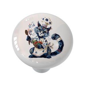 May include: White ceramic cabinet knob featuring a whimsical illustration of a blue cat chef. The cat wears a chef's hat and apron, holding a wooden spoon and a basket of vegetables. The design incorporates floral and folk art elements.