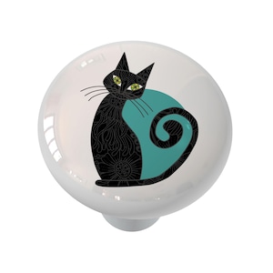 May include: A white ceramic cabinet knob featuring a black cat design. The cat has intricate floral patterns and green eyes, set against a teal blue backdrop. The knob is round with a silver-tone base.