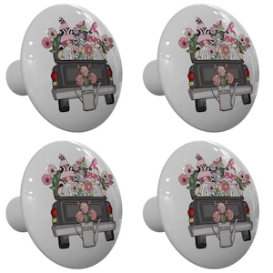 May include: Four white ceramic knobs with a black pickup truck carrying two gnomes with pink and white hats and flowers in the back. The truck has a watering can in the back.
