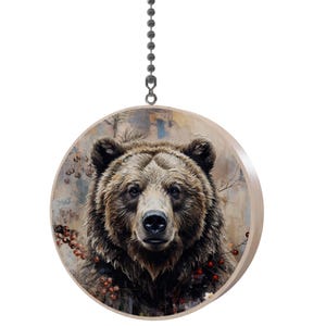 May include: Round wooden ornament featuring a detailed illustration of a brown bear's face. The bear has dark eyes and a furry coat. The background has a muted, painterly style with hints of red berries and branches. A metal chain is attached for hanging.