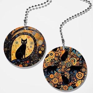 May include: Two circular pendants hanging from silver ball chains. The left pendant features a single black cat centered against a yellow and blue abstract background. The right pendant displays a repeating pattern of black cats amidst orange and blue circles.