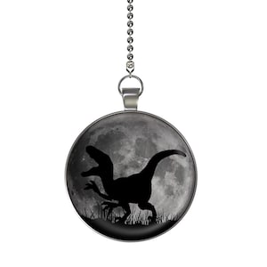 May include: A silver ceiling fan pull chain with a round, black and white pendant featuring a silhouette of a velociraptor in front of a moon.