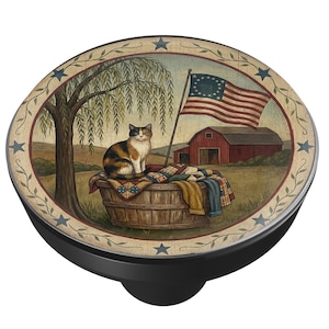 May include: A decorative cabinet knob featuring a pastoral scene with a calico cat sitting on a basket of quilts. The image includes a red barn, American flag, and a weeping willow tree. The knob has a black base and an oval design.