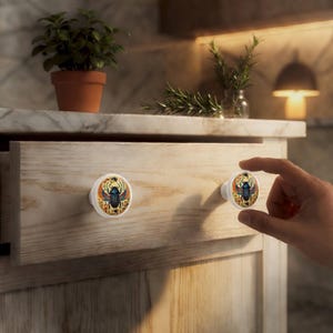 Puede incluir: Pomos de armario blancos con un dise&ntilde;o de escarabajo egipcio. Los pomos presentan un escarabajo azul con alas naranjas y s&iacute;mbolos jerogl&iacute;ficos sobre un fondo crema. Los pomos est&aacute;n fijados a un caj&oacute;n de madera clara.