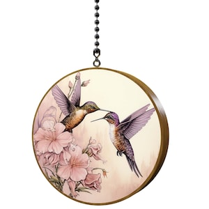 May include: A round wooden wall hanging with a beige background featuring two hummingbirds in flight. The hummingbirds are painted in shades of purple, brown, and green. The background features pink flowers and a soft, watercolor-like effect.