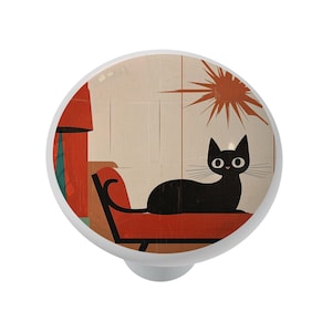 May include: A white cabinet knob with a retro illustration of a black cat lounging on a red chair. The cat has large, round eyes and long whiskers. The background features a window and a sunburst design.