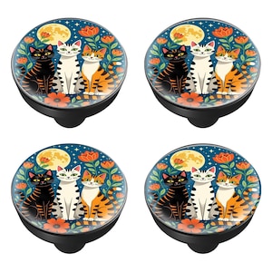 May include: Four black cabinet knobs with a colorful cat design. Each knob features a black cat, a white cat, and an orange tabby cat sitting together under a full moon and flowers on a dark blue background.