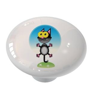 May include: White ceramic cabinet knob with a cartoon cat design. The cat is black with yellow eyes and pink accents, set against a blue sky and green grass background. The knob is round and glossy.