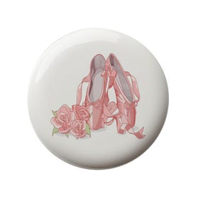 May include: A pair of pink ballet shoes with ribbons tied together, resting on a white background with three pink roses.